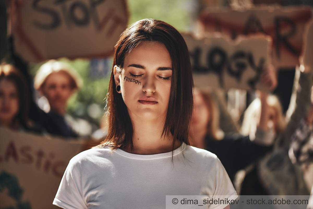 Eine junge Frau mit geschlossenen Augen trägt das Wort "FREEDOM" auf ihrem Gesicht. Im Hintergrund sind weitere Personen zu sehen, die Plakate halten. Die Szene vermittelt eine Botschaft des Protestes und des Strebens nach Freiheit.