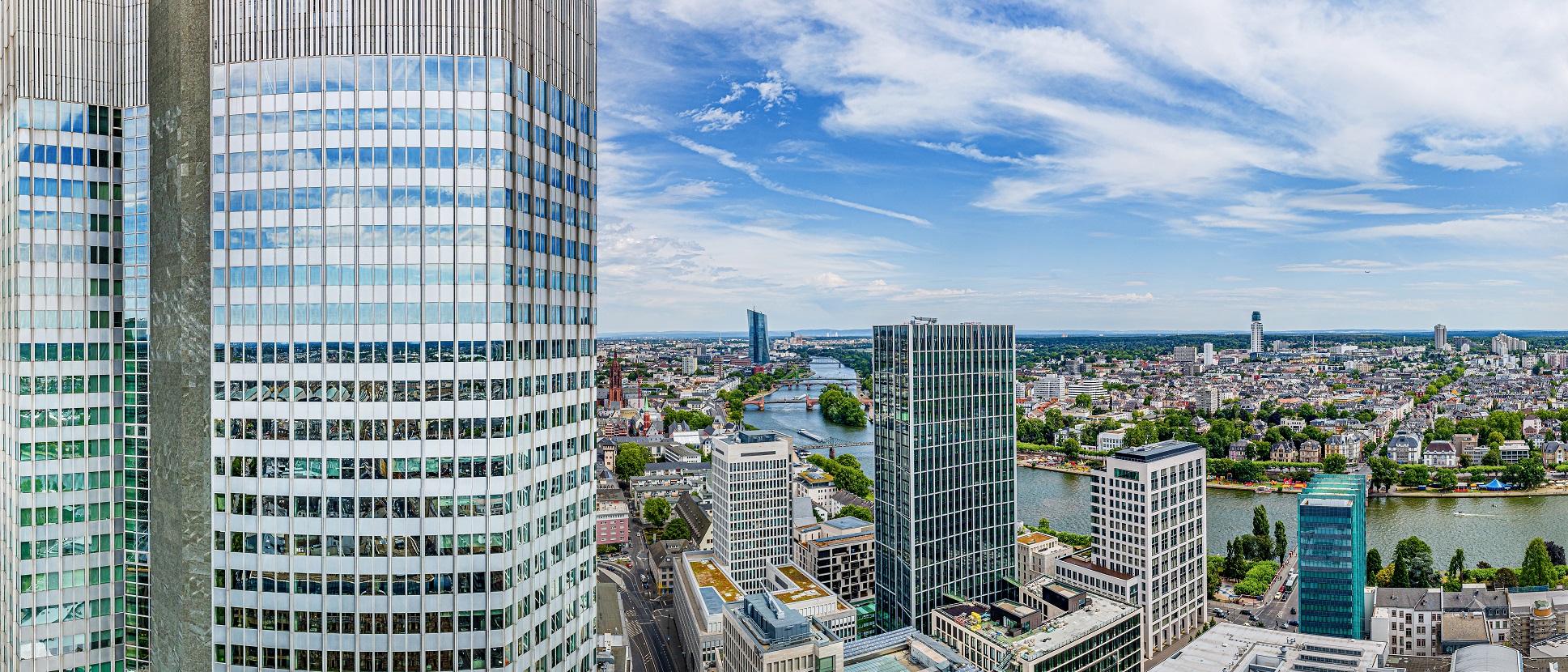 Blick auf eine Stadtlandschaft mit modernen Wolkenkratzern, einem Fluss und einem blauen Himmel. Die Architektur ist vielfältig, mit Beton- und Glasgebäuden. Im Hintergrund sind Bäume und einige historische Gebäude zu sehen.
