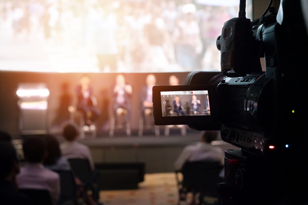 Eine Kamera filmt eine Podiumsdiskussion, auf der mehrere Personen sitzen. Im Hintergrund ist ein unscharfes Publikum zu sehen. Die Teilnehmer scheinen lebhaft zu sprechen, während die Kamera auf sie fokussiert ist.