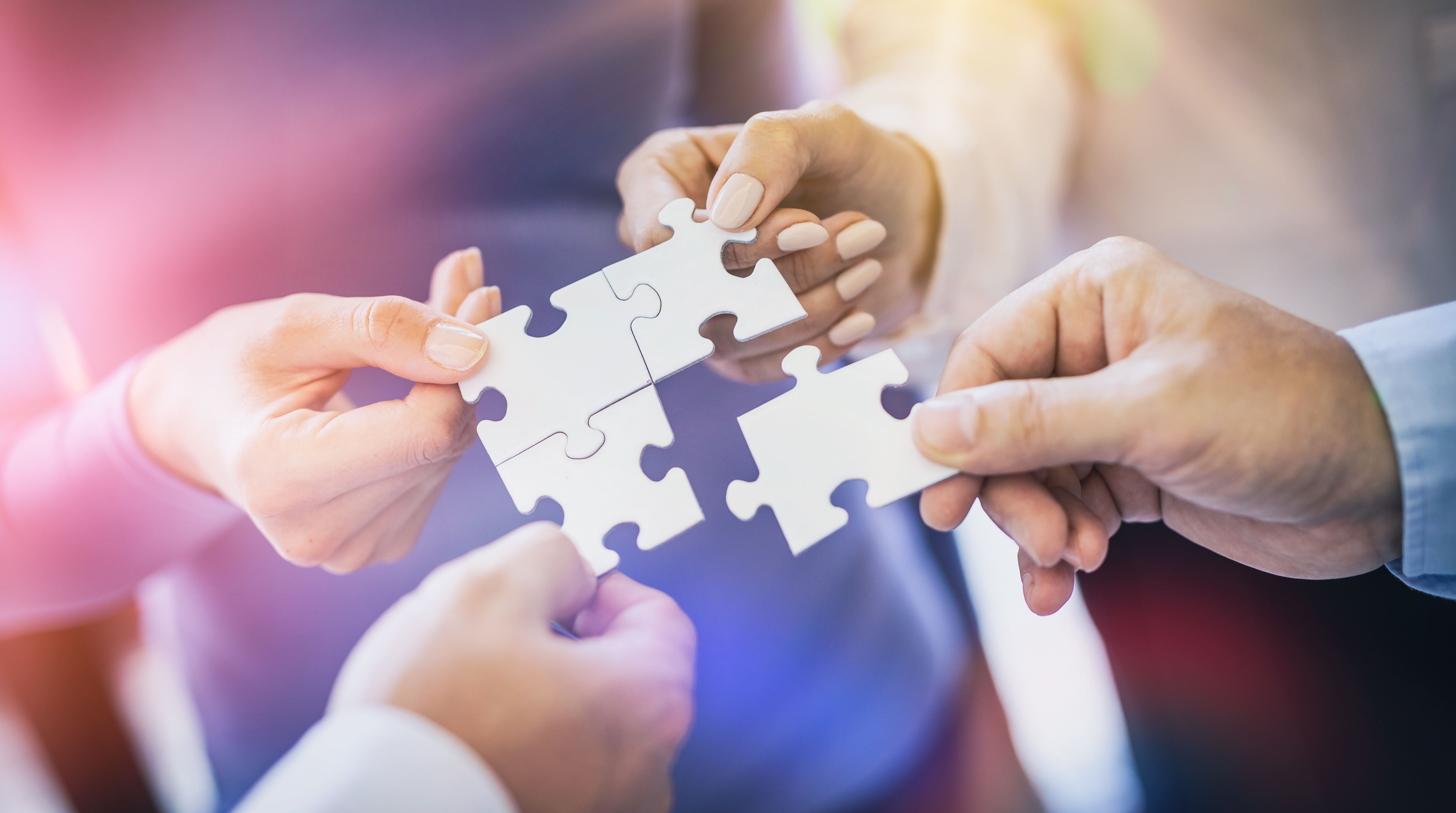 Four hands are holding puzzle pieces, working together to fit them together. The image represents collaboration, teamwork, and the idea of solving problems or completing a task collectively.