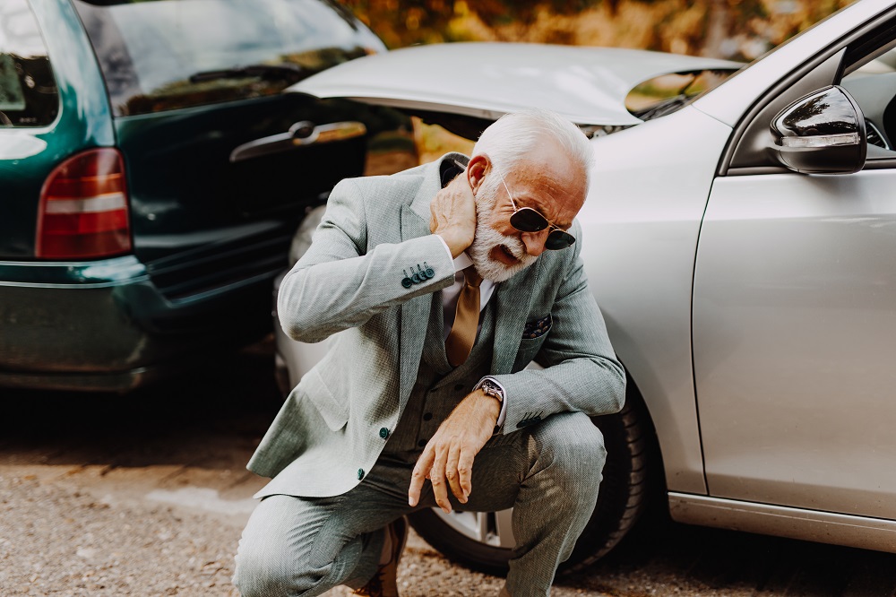 Ein älterer Mann in einem eleganten grauen Anzug und Sonnenbrille kniet an einem Auto. Er hält sich die Hand an den Nacken und sieht besorgt aus. Im Hintergrund steht ein weiteres Fahrzeug.