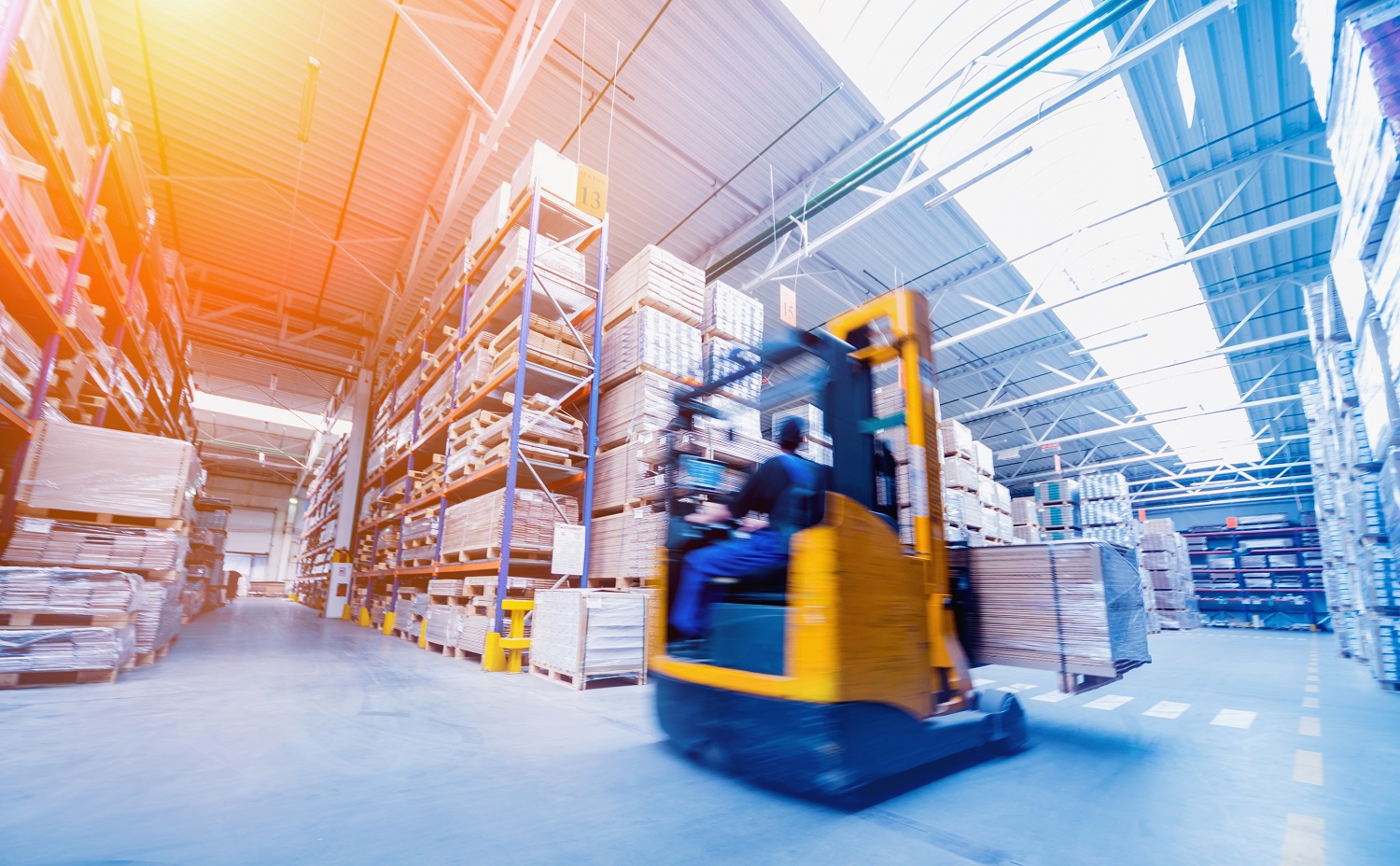 A forklift is moving down a wide aisle in a warehouse filled with tall shelves stacked with pallets of goods. The environment is bright and spacious, with numerous items organized neatly along the sides.