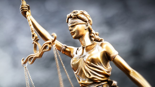 A bronze statue of Lady Justice, blindfolded, holds a set of scales in one hand and a sword in the other. The blindfold symbolizes impartiality, while the scales represent the weighing of evidence, and the sword signifies the authority of the law.