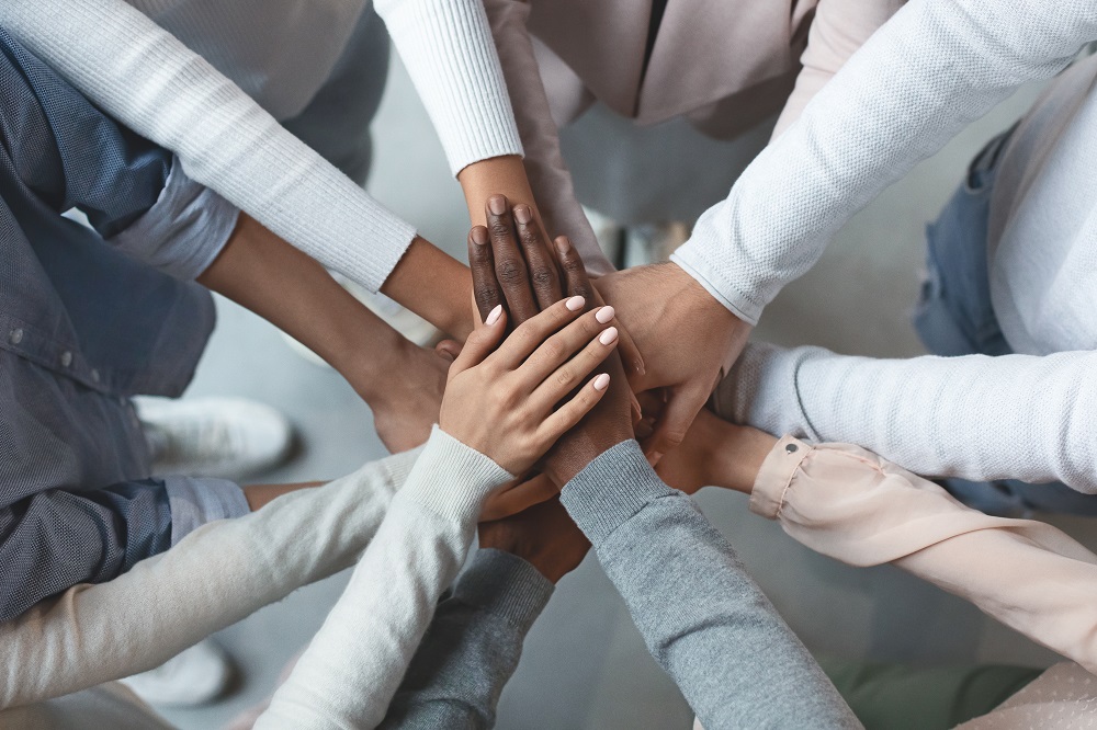 A diverse group of hands is stacked together in a show of unity and teamwork. The hands vary in skin tones and styles, symbolizing collaboration and support among individuals from different backgrounds.
