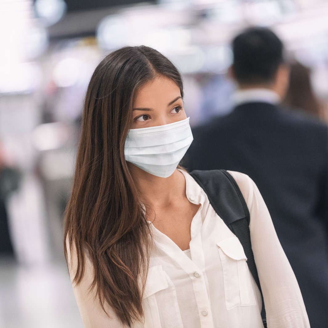 Eine junge Frau mit langen, braunen Haaren trägt eine medizinische Maske und ein weißes Hemd. Sie schaut nachdenklich in eine unbestimmte Richtung, während im Hintergrund unscharfe Silhouetten von anderen Menschen zu sehen sind.