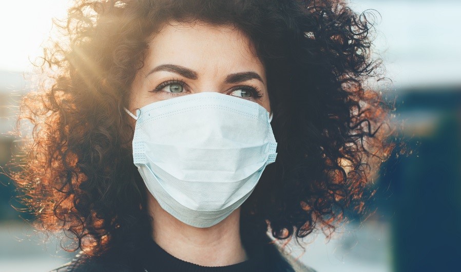 Eine Person mit lockigem, braunem Haar trägt eine medizinische Maske. Der Blick ist nachdenklich, während sanftes Licht sie umgibt. Die Umgebung ist unscharf, was den Fokus auf das Gesicht und die Maske legt.