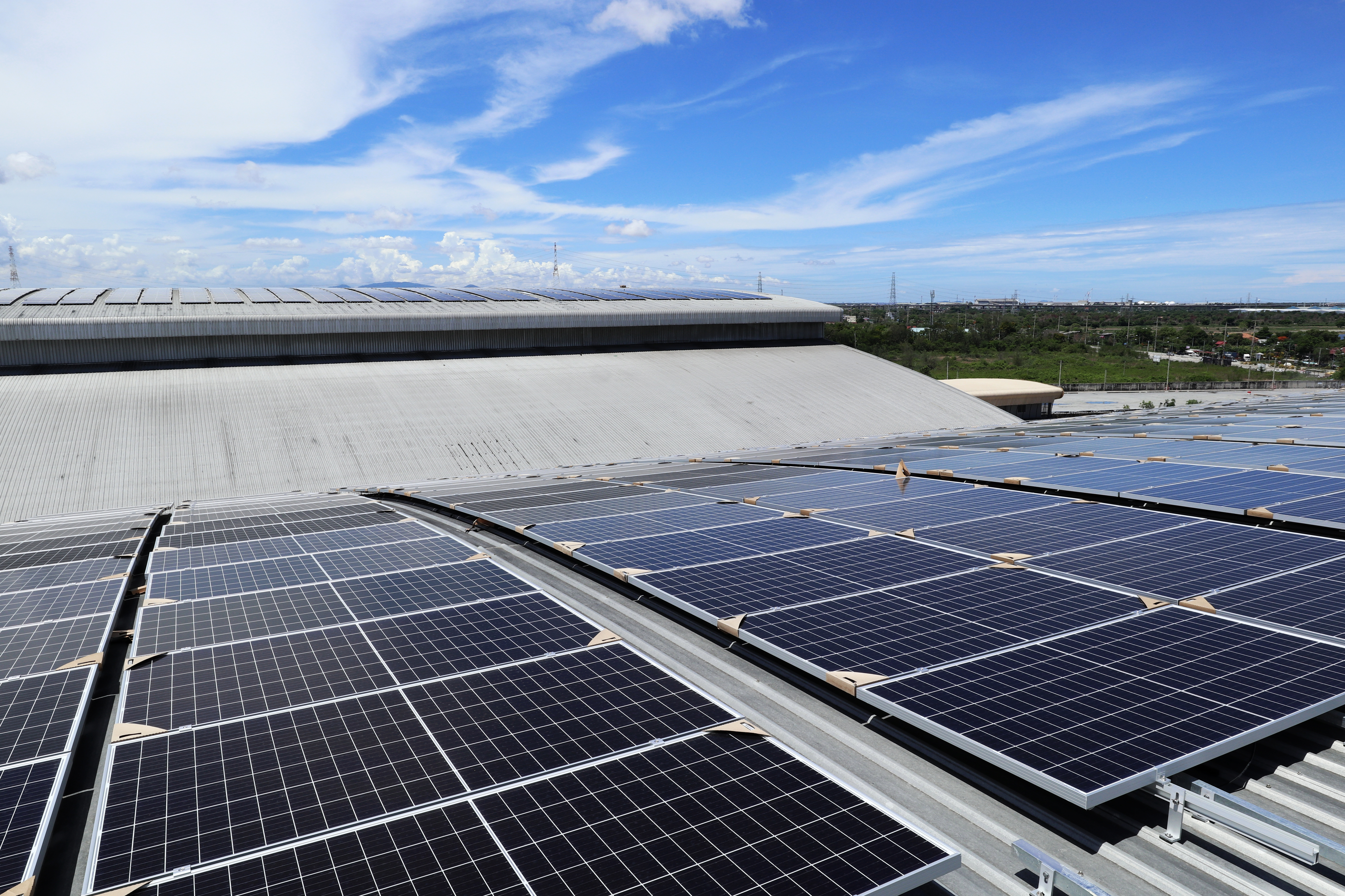 A rooftop covered with solar panels is shown under a clear blue sky. The panels are arranged in rows, reflecting sunlight, while the building's structure features a sloped roof. Green landscape can be seen in the background, emphasizing the renewable energy focus.