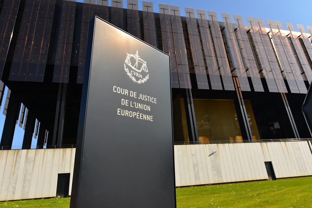 Ein schwarzes Schild steht vor einem modernen Gebäude. Auf dem Schild steht in goldenen Buchstaben „COUR DE JUSTICE DE L'UNION EUROPÉENNE“ und das Symbol einer Waage. Das Gebäude hat große, glasähnliche Paneele und eine helle, glatt gestaltete Fassade.