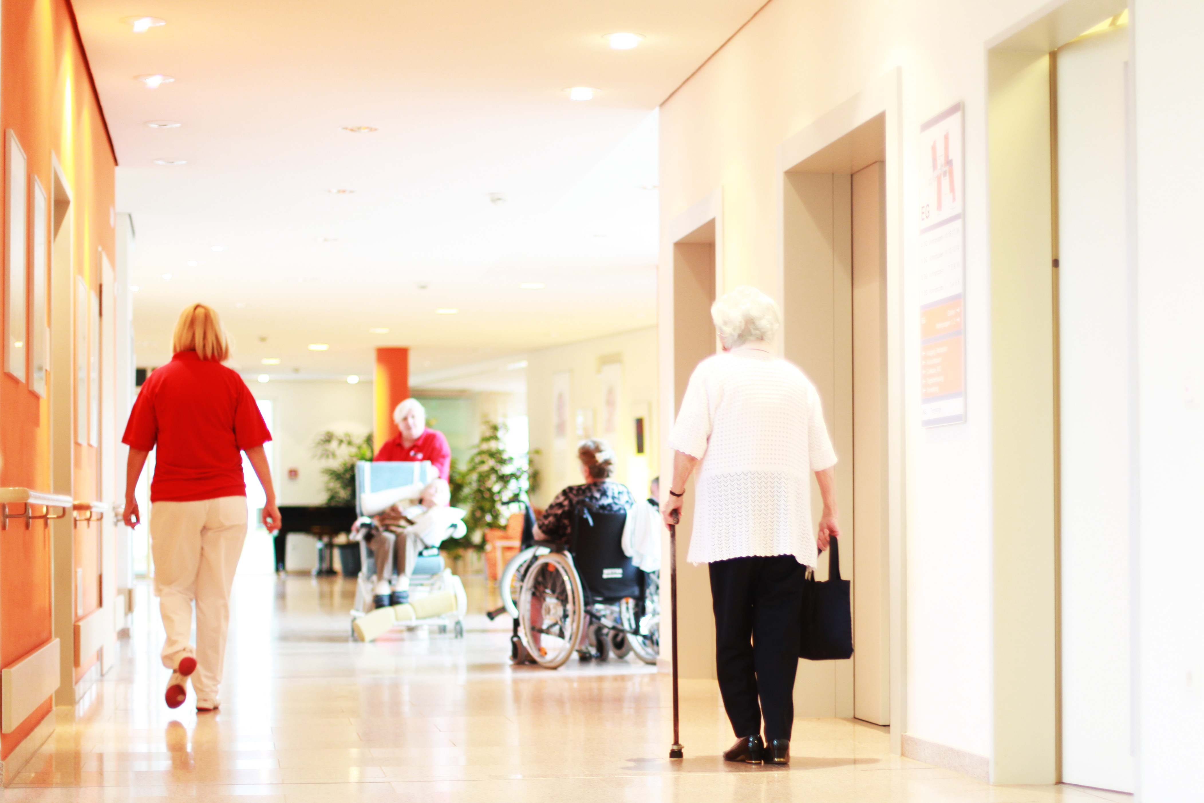 A well-lit corridor in a care facility features, on one side, a staff member in red walking towards two elderly individuals, one with a cane and another in a wheelchair. The scene conveys a supportive environment for residents.