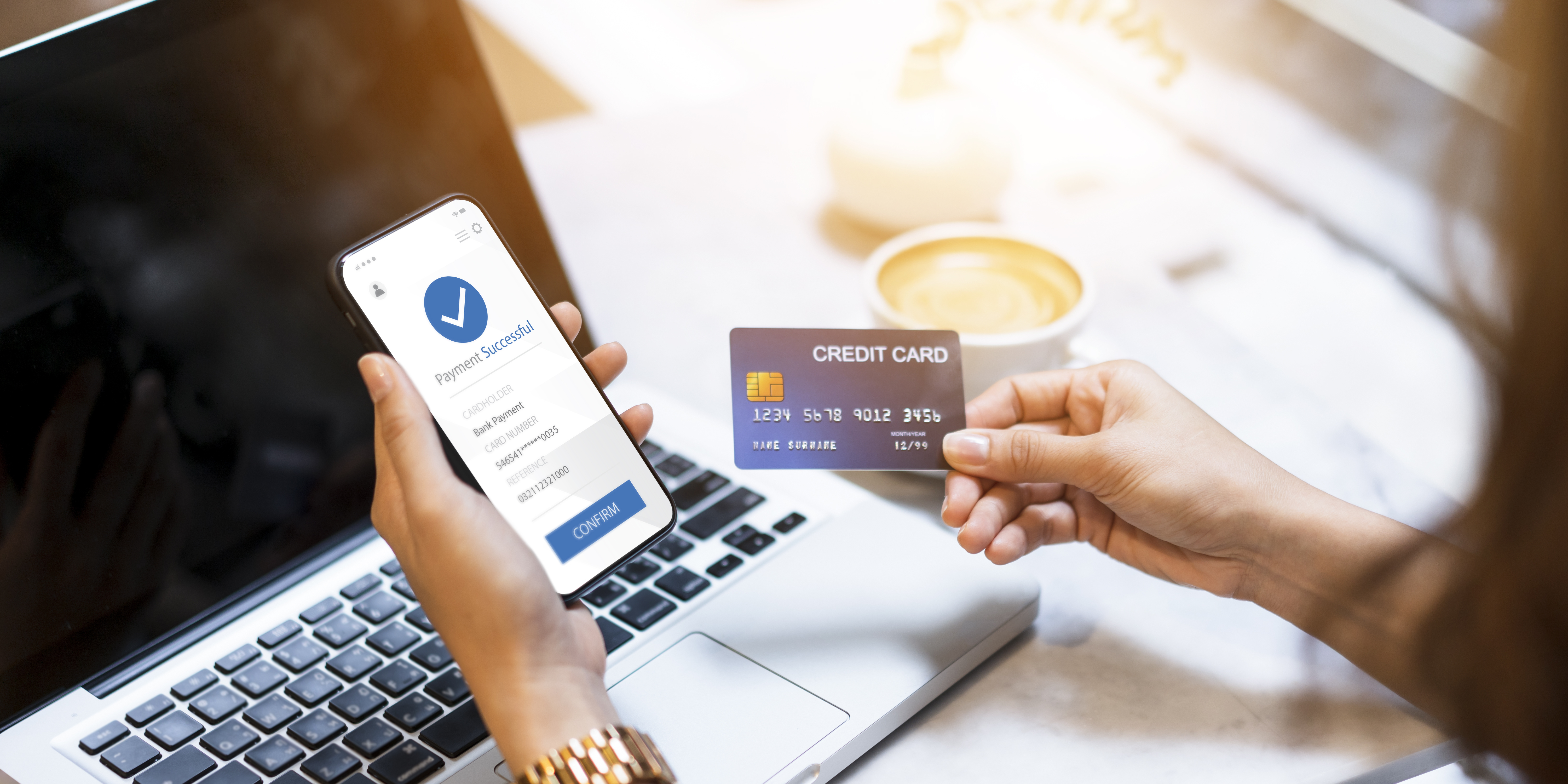 A person holds a smartphone displaying a payment confirmation screen while also holding a credit card. A laptop is visible in the background, and a cup of coffee is on the table. The scene suggests online shopping or payment processing.