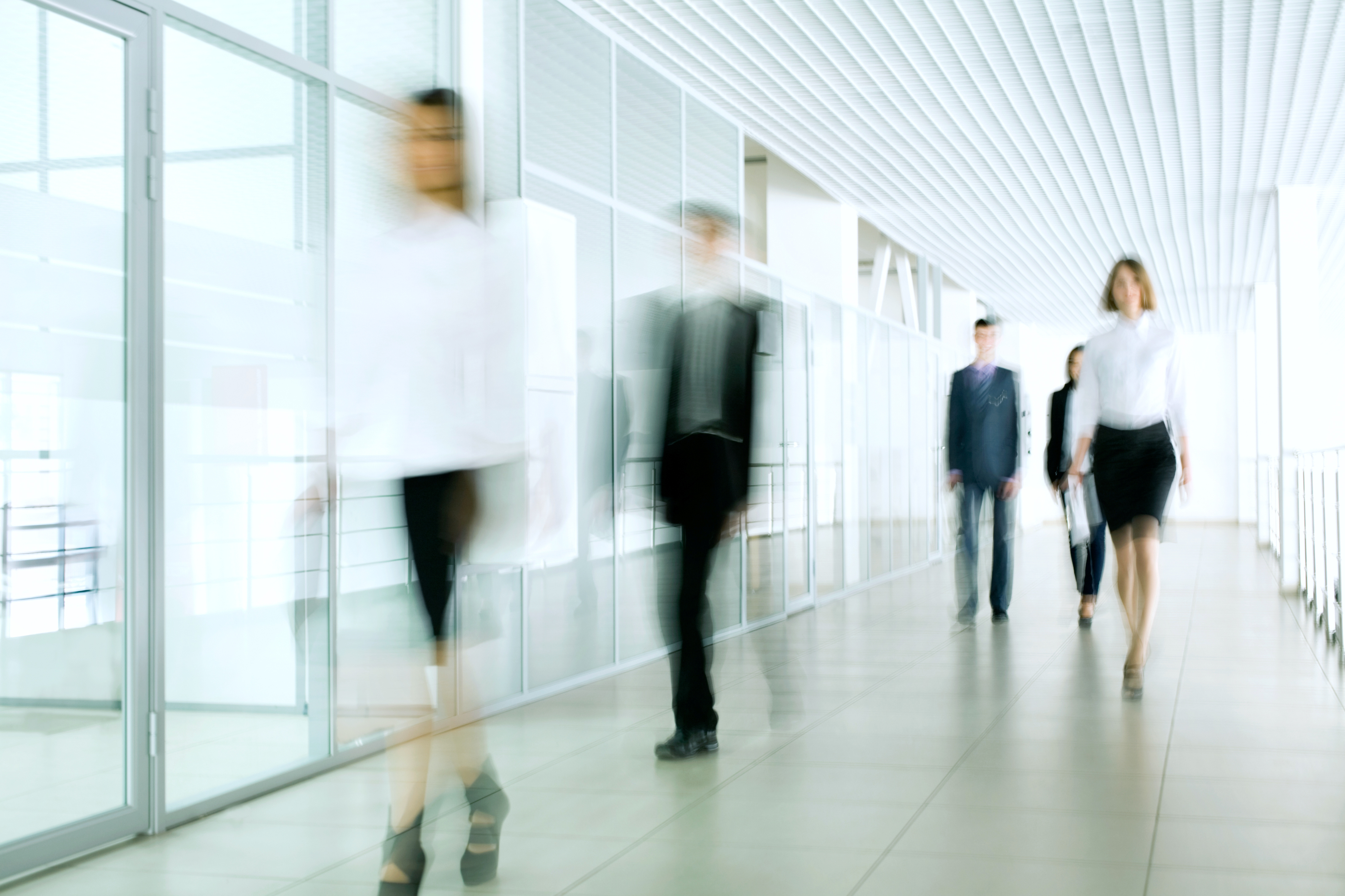A group of business professionals walks through a modern, brightly lit hallway. The figures appear slightly blurred, indicating movement. The setting suggests a corporate environment, with glass walls and a clean, minimalist design.