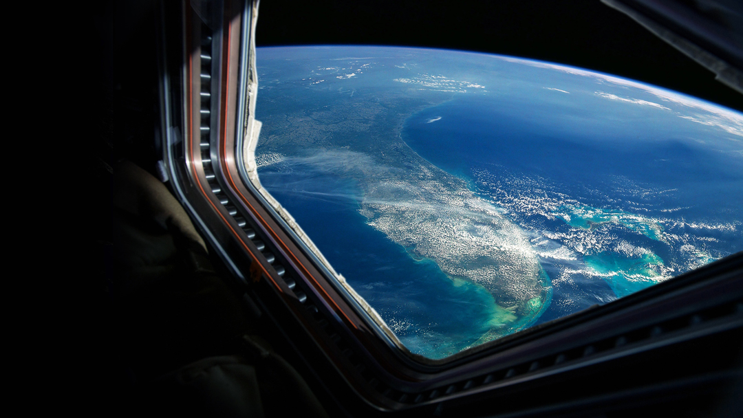 Blick auf die Erde aus einem Raumschifffenster, das den Ozean, Wolken und Küstenlinien zeigt. Die Farben des Wassers variieren zwischen tiefblau und grünlich, während weiße Wolken die Atmosphäre überziehen. Der Blick vermittelt ein Gefühl von Weite und der Schönheit des Planeten.