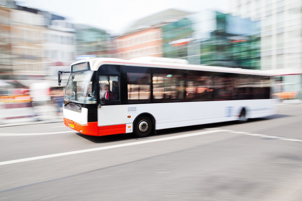 Ein schneller, moderner Bus mit weißen und orangen Farben fährt auf einer Stadtstraße. Hintergrund unscharf, um Bewegung darzustellen.
