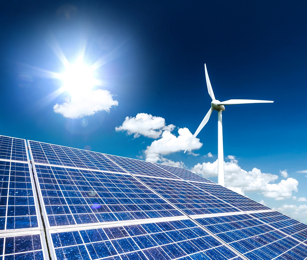 A bright sun shines over a field of solar panels in the foreground, with a wind turbine standing tall in the background. The sky is blue with scattered clouds, symbolizing renewable energy and sustainability.