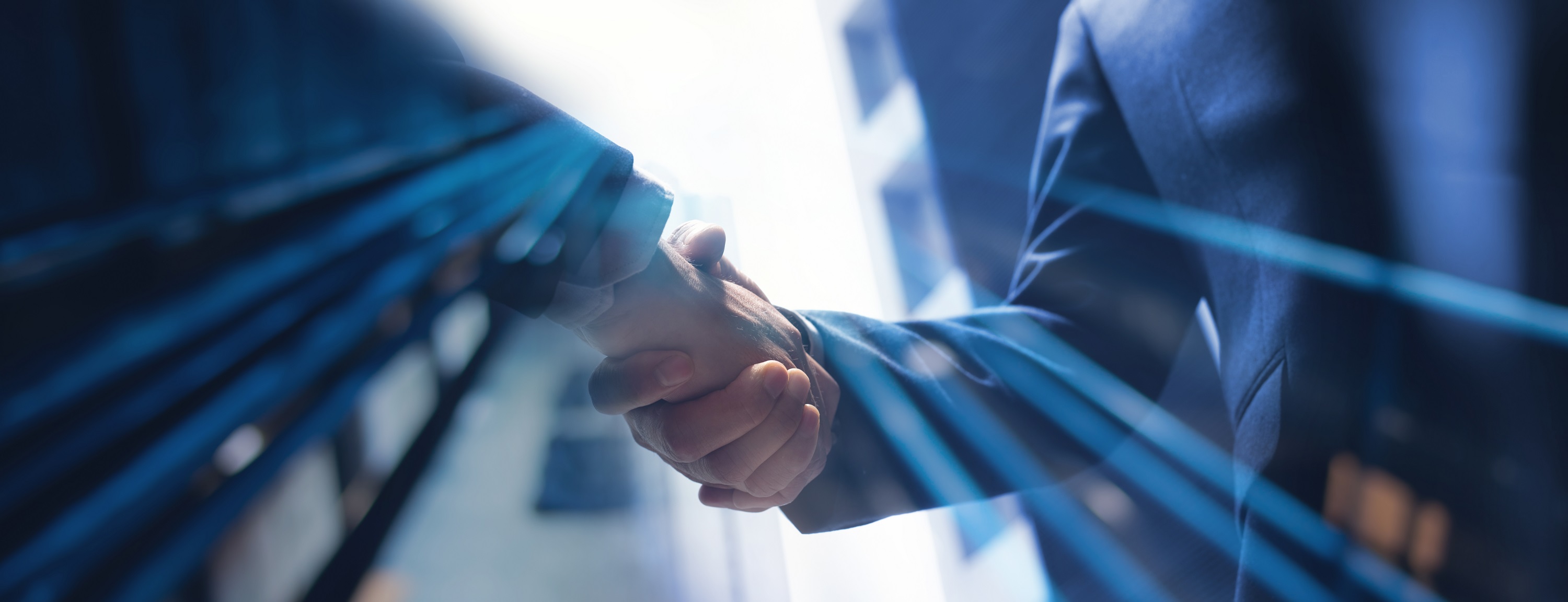 Two hands are shaken in a business setting, symbolizing partnership and agreement. The focus is on the handshake, with a blurred background that suggests a professional atmosphere. Light rays are visible, adding emphasis to the moment of connection.