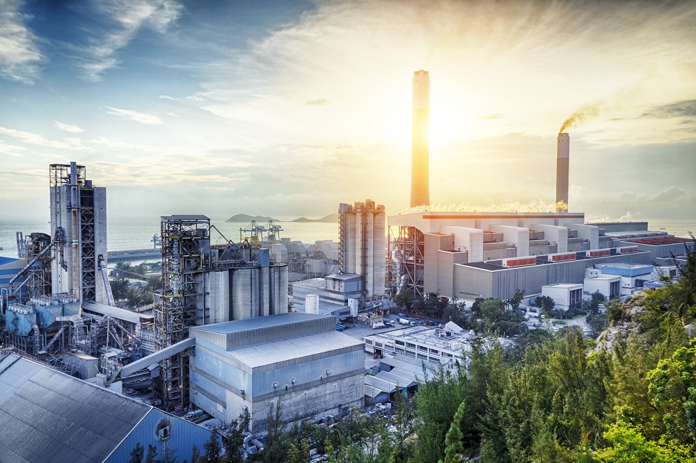 A large industrial power plant is depicted, featuring tall smokestacks emitting smoke. The plant is situated near a body of water with mountains in the background, and the sun is rising, casting a warm glow over the scene.