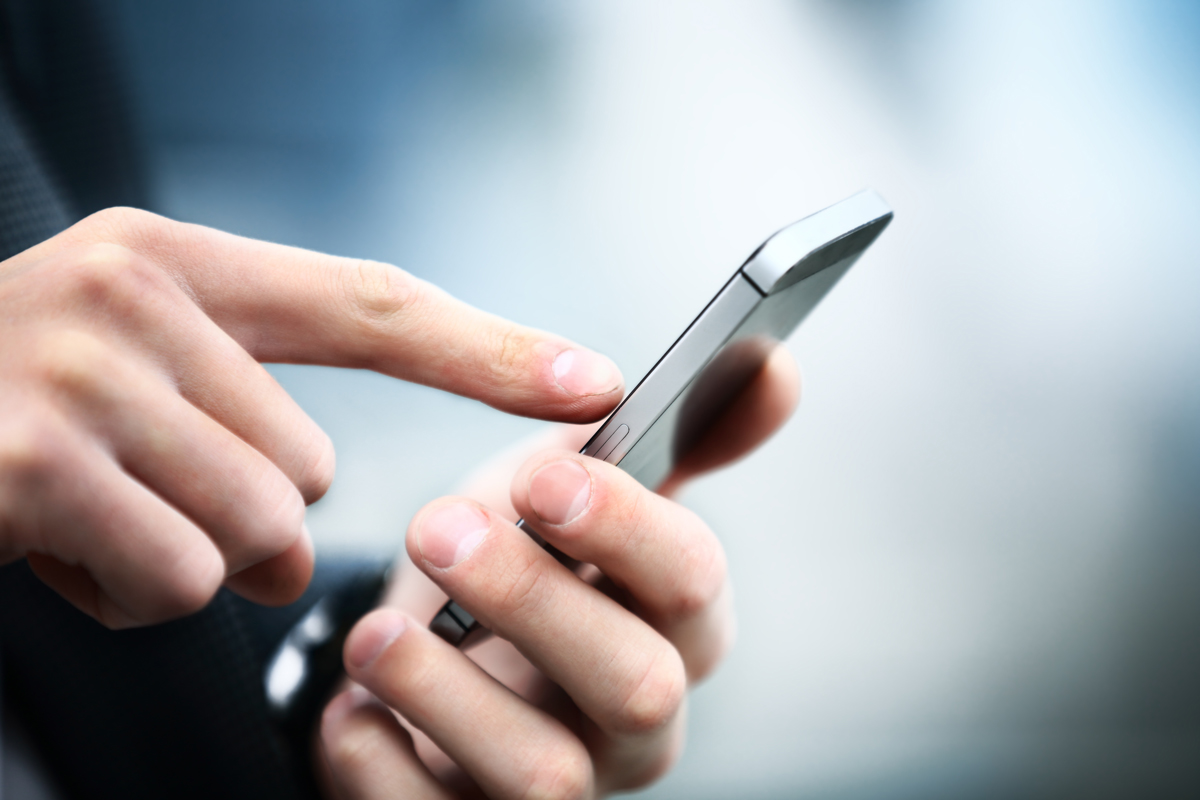 Eine Hand hält ein Smartphone, während ein Finger auf dem Bildschirm tippt. Die Umgebung ist unscharf, was den Fokus auf die Interaktion mit dem Gerät legt.