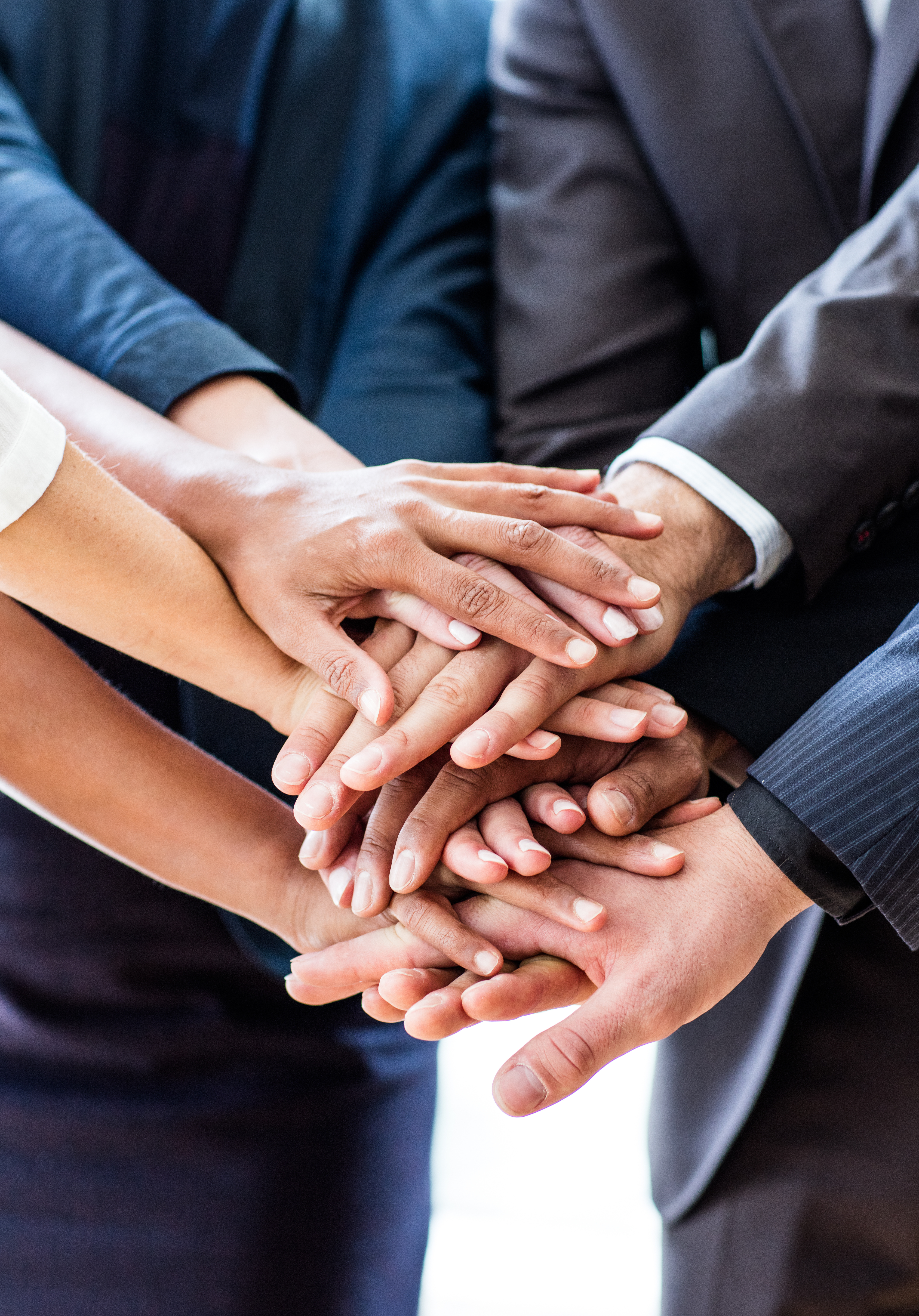 A group of diverse hands are stacked on top of each other, symbolizing teamwork and unity. The hands vary in skin tone and are well-groomed, highlighting collaboration among individuals from different backgrounds.