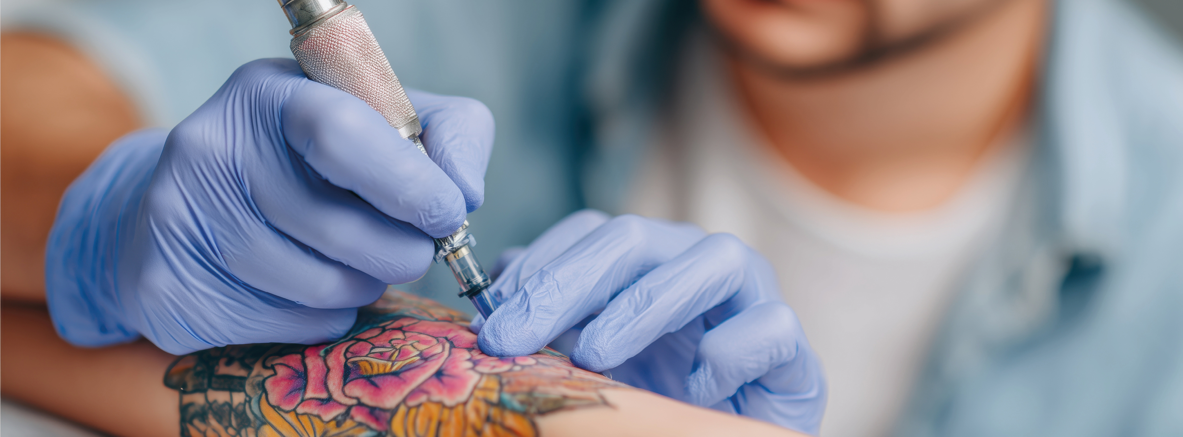 Ein Tätowierer trägt mit einer Maschine ein farbenfrohes Tattoo einer Blume auf den Unterarm einer Person auf. Die Hände des Tätowierers sind mit blauen Handschuhen geschützt.