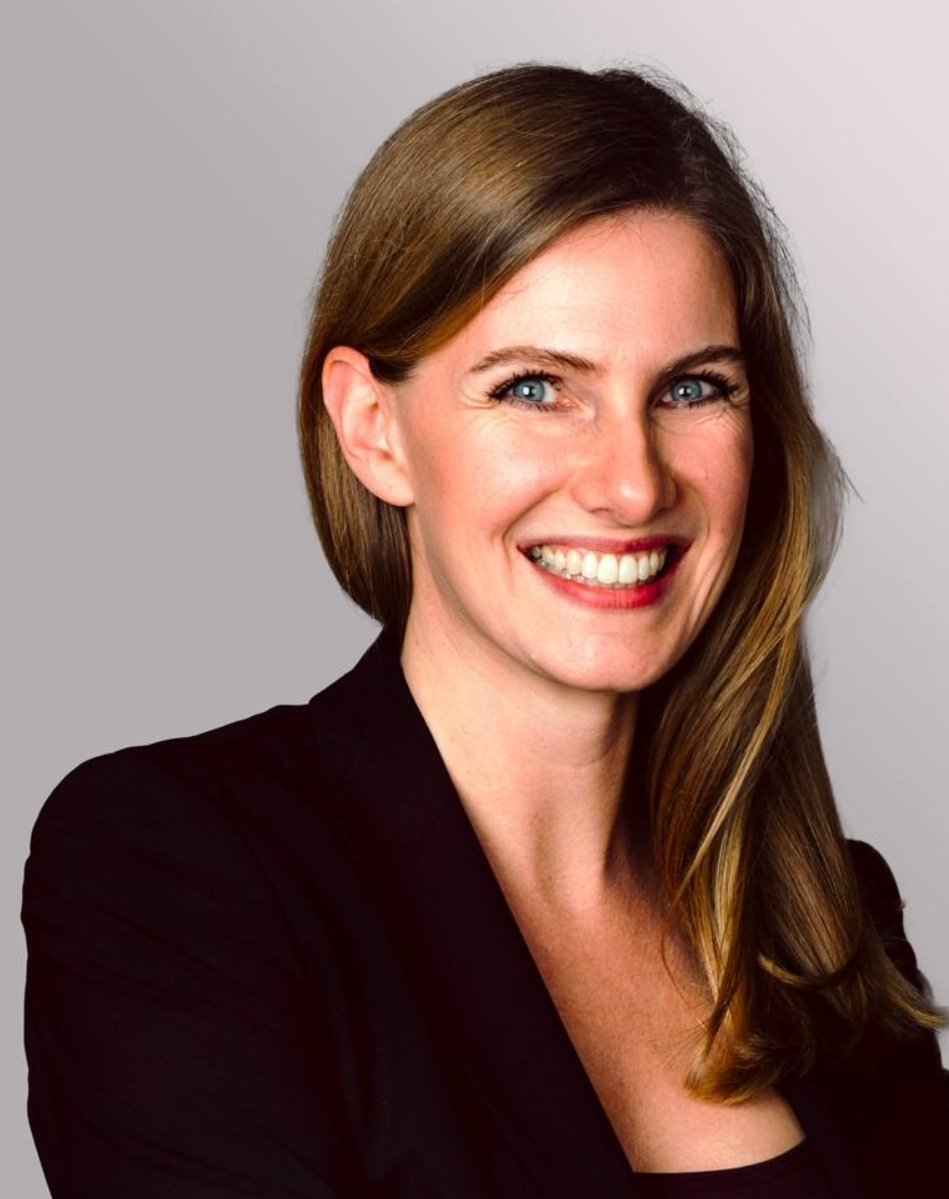 A woman with long, straight brown hair smiles warmly, wearing a black blazer. Her blue eyes are bright and engaging, conveying a friendly demeanor against a soft gradient gray background.