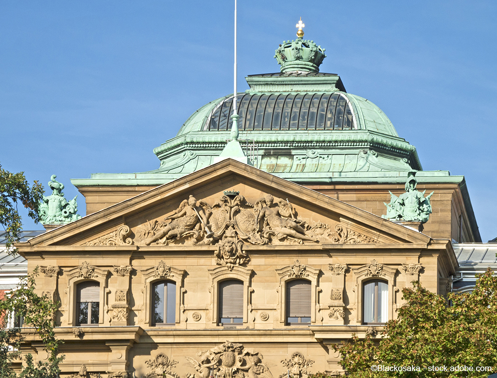 Die obere Fassade eines historischen Gebäudes mit einer grünen Kuppel und dekorativen Reliefs. In der Mitte befindet sich ein Wappen, flankiert von weiteren kunstvollen Details. Der Himmel ist blau und klar, was dem Gebäude eine majestätische Präsenz verleiht.