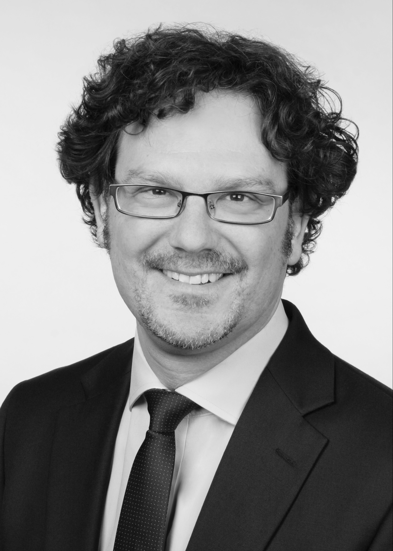 A man with curly hair and glasses smiles in a formal suit and tie. The background is plain, enhancing the focus on his friendly expression.