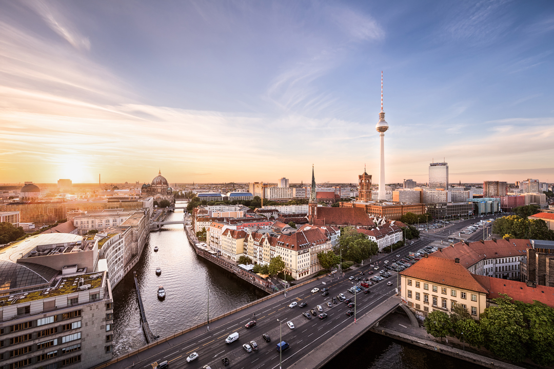 A panoramic view of Berlin at sunset, showcasing the River Spree winding through the city. The iconic Fernsehturm (TV tower) stands tall among a mix of historic and modern buildings, while bustling streets and greenery add vibrancy to the urban landscape.