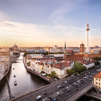 Ein Blick auf Berlin zur Dämmerung, mit der Spree im Vordergrund und historischen Gebäuden entlang des Ufers. Im Hintergrund ist der Fernsehturm sichtbar, umgeben von moderner Architektur und einer belebten Straße. Der Himmel zeigt sanfte Farben des Sonnenuntergangs.