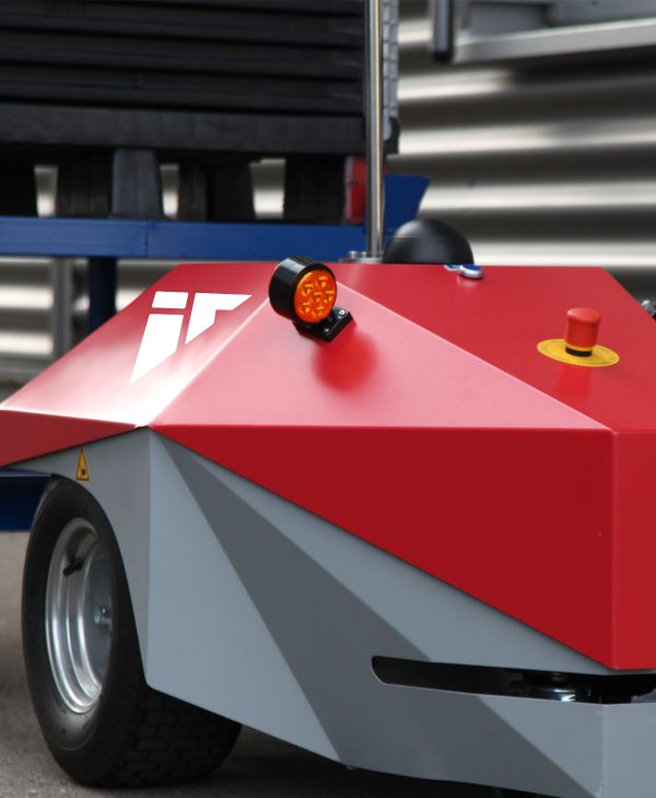 A close-up view of a small, angular, red and gray automated vehicle. It features an orange flashing light on top and a yellow button, designed for operational use in industrial or warehouse settings. The background is blurred, focusing on the vehicle's details.