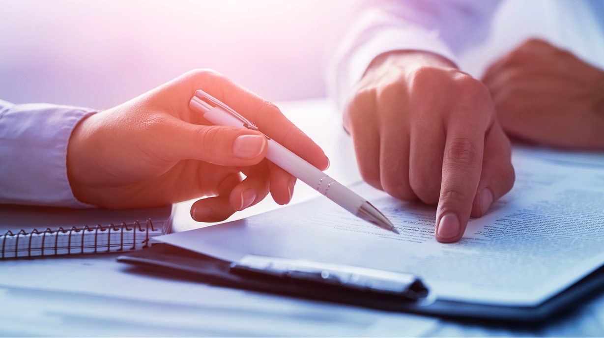 Zwei Hände auf einem Tisch, eine hält einen Stift und zeigt auf ein Dokument, während die andere Hand auf demselben Dokument zeigt. Im Hintergrund sind ein Notizbuch und ein weiterer Stift zu sehen.