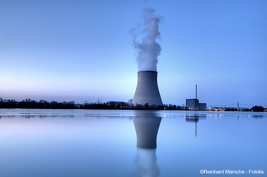 Ein Kühlturm eines Kernkraftwerks steht am Ufer eines ruhigen Gewässers, während Dampfwolken aus seiner Spitze aufsteigen. Die Umgebung ist in sanfte Blautöne gehüllt, und die Spiegelung des Turms ist im Wasser sichtbar.