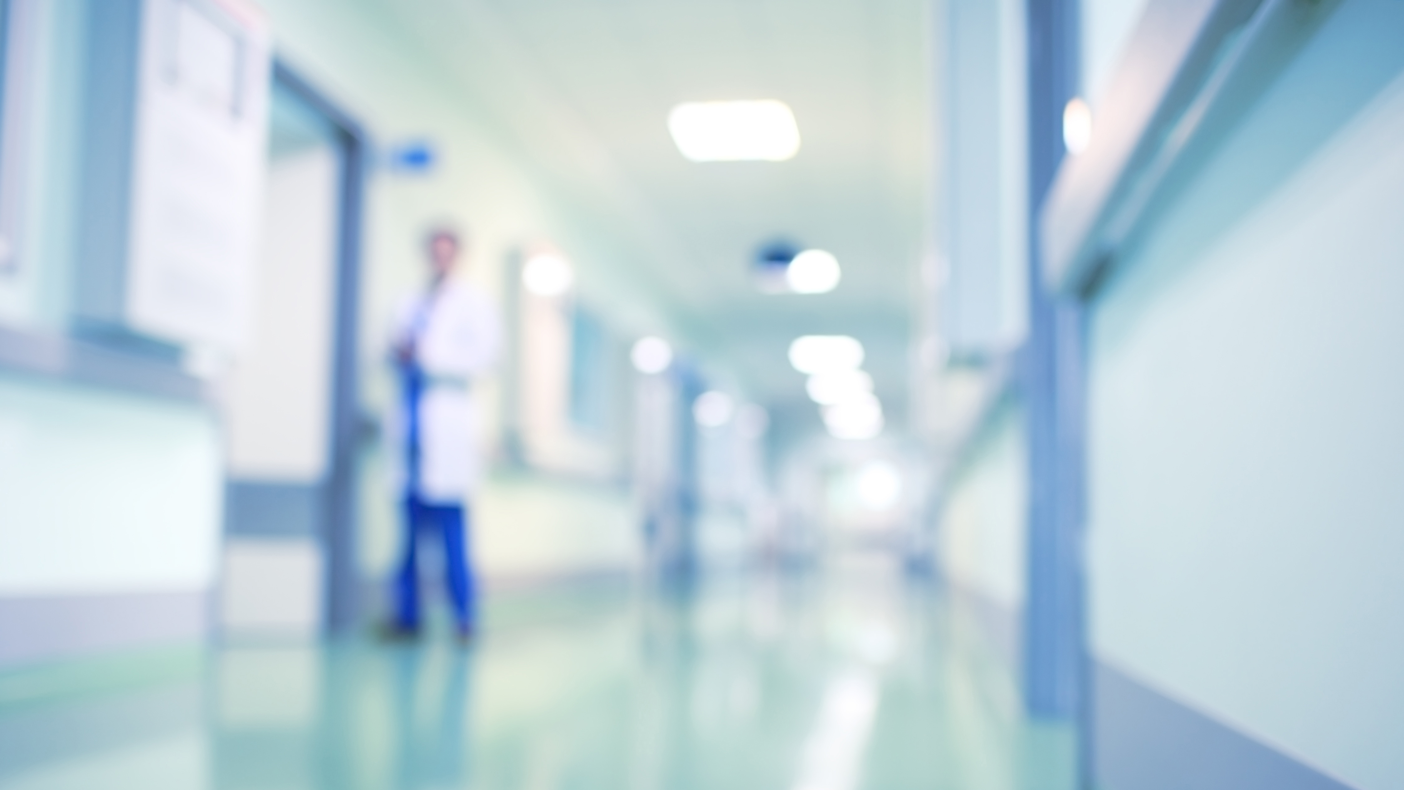 Ein unscharfer Krankenhausflur mit einem Arzt, der im Hintergrund steht. Der Flur ist hell erleuchtet und zeigt Krankenhauszimmer auf beiden Seiten. Der Fokus liegt auf der Präsenz des Arztes im Raum.