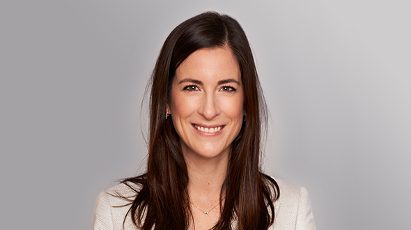 A woman with long, dark hair smiles warmly at the camera. She is wearing a light-colored blazer and a delicate necklace, set against a neutral gray background. Her expression conveys confidence and approachability.