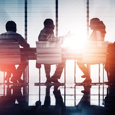 Die Silhouette von mehreren Personen in einem Meetingraum, die an einem Tisch sitzen. Eine Person ist aktiv im Gespräch, während die anderen zuhören. Sanftes Licht strömt durch große Fenster, was eine dynamische und kollegiale Atmosphäre schafft.