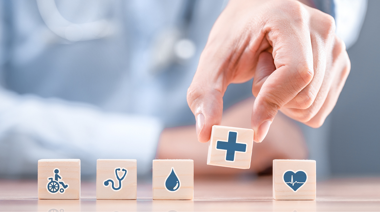 Eine Hand platziert einen Würfel mit einem Kreuzsymbol auf einem Tisch. Vor der Hand liegen vier weitere Würfel mit Symbolen für Gesundheit, Wasser, einen Stethoskop und einen Rollstuhl. Die Szene vermittelt das Thema Gesundheit und medizinische Versorgung.