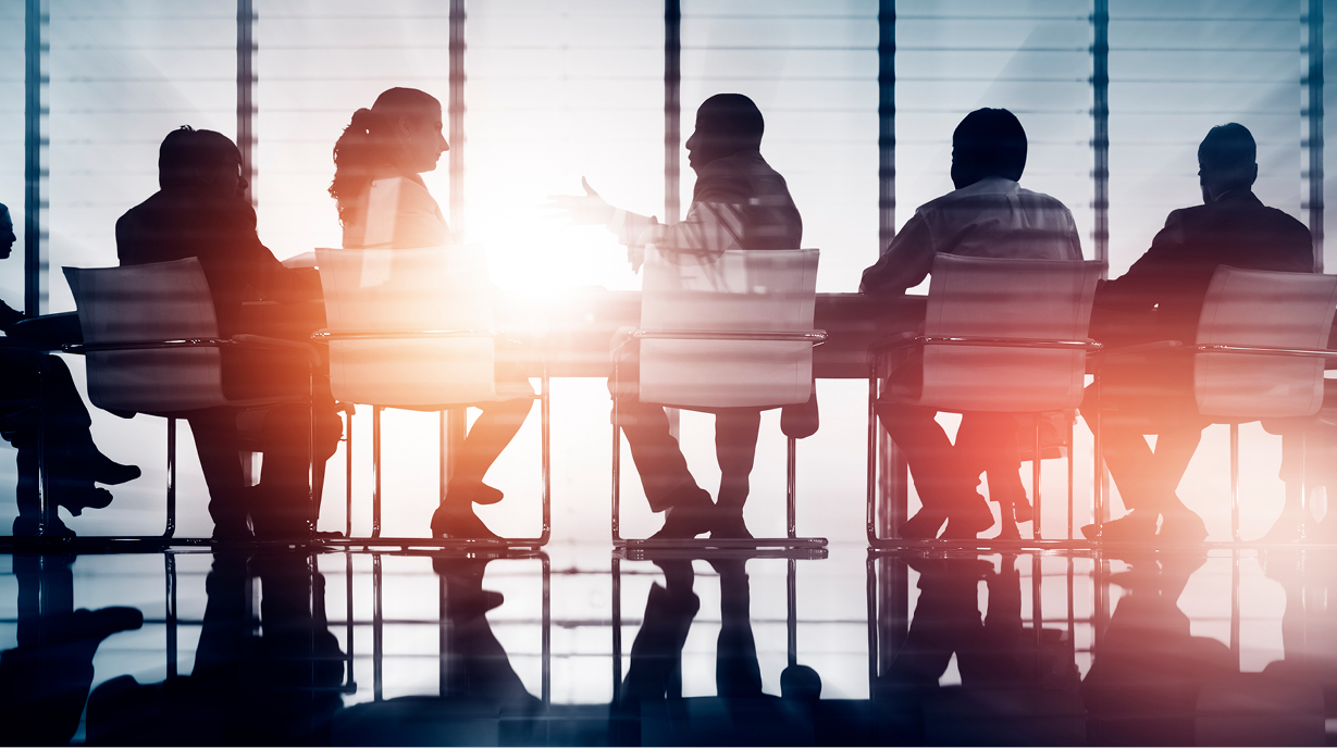 Silhouetten von fünf Personen, die an einem Konferenztisch sitzen. Eine Person steht und spricht, während die anderen zuhören. Im Hintergrund strömt Sonnenlicht durch große Fenster, was eine helle Atmosphäre schafft.