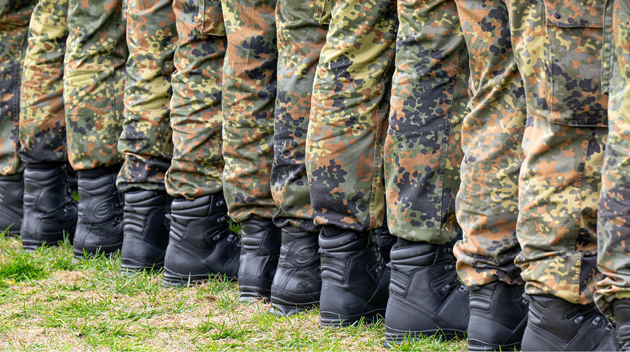 Reihe von Beinen in Uniform, bestehend aus militärischer Tarnkleidung und schwarzen Stiefeln, die auf einer Grasfläche aufgestellt sind. Die Hosen sind aus verschiedenen Tarnmustern gefertigt und stehen in gerader Linie.