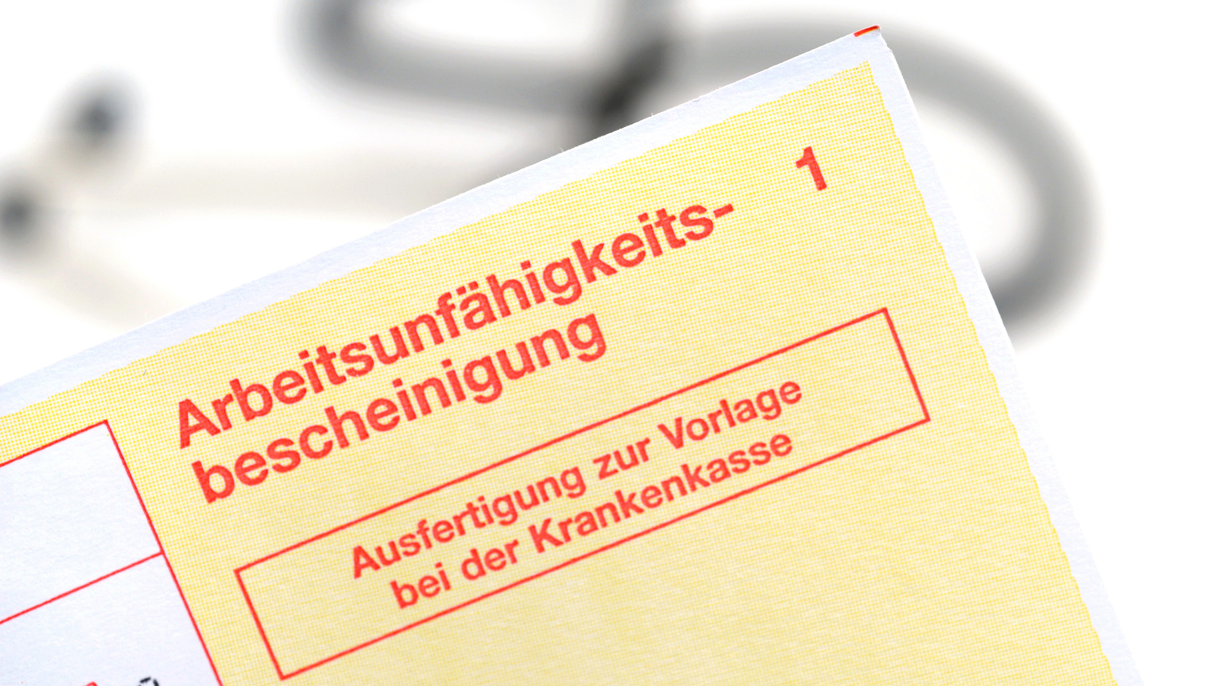 Ein gelbes Formular mit dem Titel "Arbeitsunfähigkeitsbescheinigung" in roter Schrift. Darunter steht "Ausfertigung zur Vorlage bei der Krankenkasse". Im Hintergrund ist eine verschwommene Darstellung eines Stethoskops zu sehen.