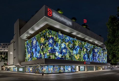 A brightly lit building at night features vibrant floral patterns covering its exterior. The building has a modern design with large windows and is marked by a prominent red sign displaying the letter "B."