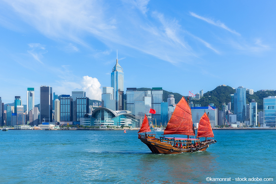 A traditional wooden junk boat with red sails sails through the water, in front of a modern Hong Kong skyline featuring towering skyscrapers and lush hills in the background. The scene reflects a blend of history and contemporary architecture under a clear blue sky.