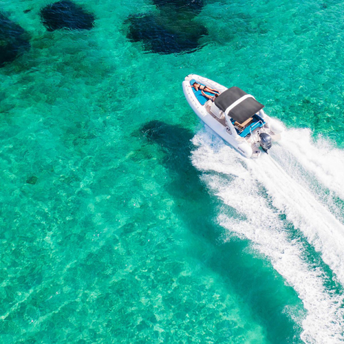 A motorboat speeds across clear turquoise waters, leaving a white wake behind. The water's surface is calm, and patches of darker areas below suggest underwater features. The scene conveys a sense of adventure and relaxation in a tropical setting.