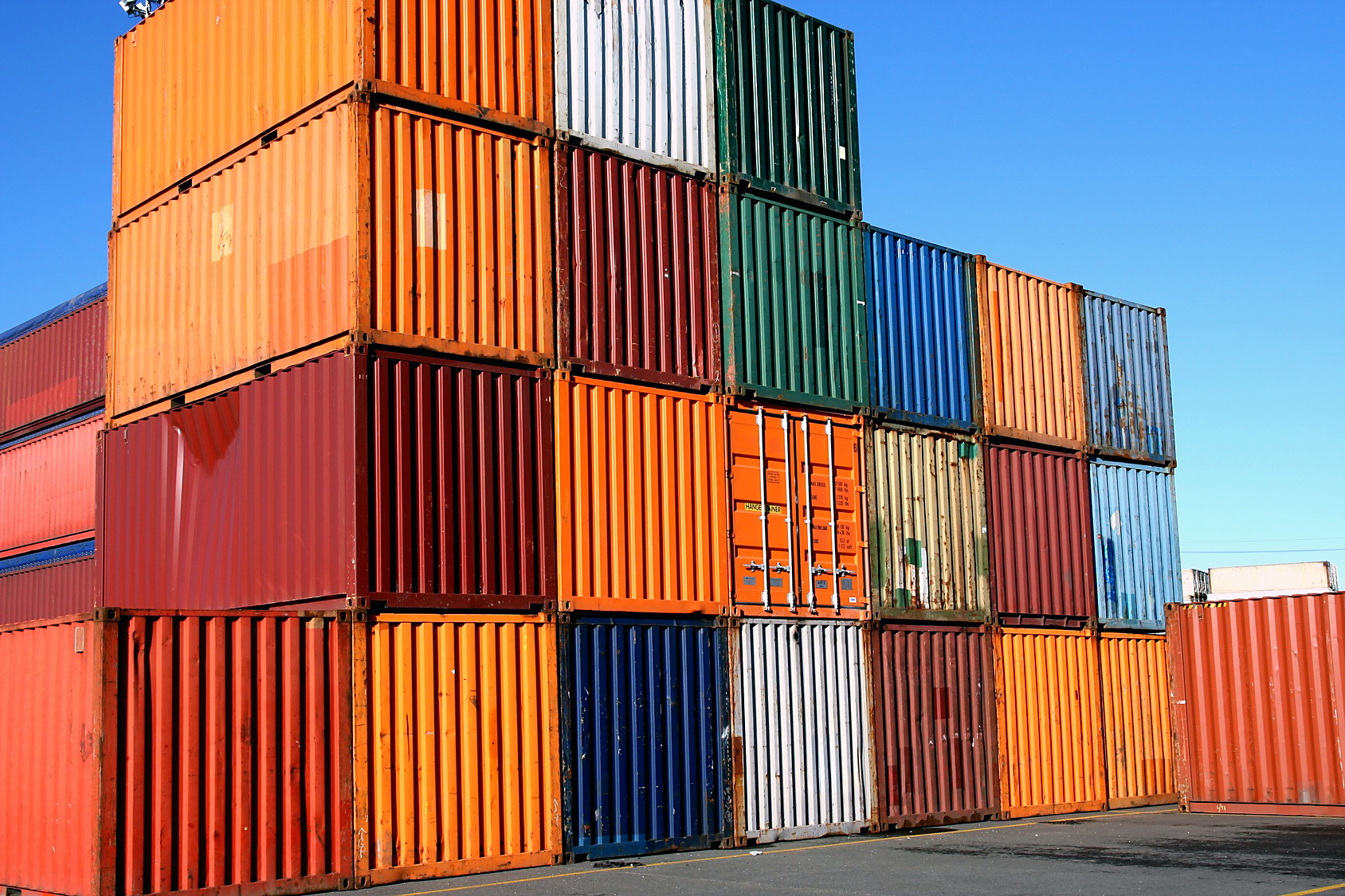 Ein Stapel von Containern in verschiedenen Farben steht aufeinander. Die Container sind aus Metall und reichen bis zu einem klaren blauen Himmel. Die Anordnung zeigt eine Mischung aus orange, grün, blau, rot und weiß.
