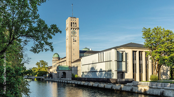 Ein klarer blauer Himmel über dem Deutschen Museum in München. Im Vordergrund fließt ein ruhiger Kanal, umgeben von Bäumen. Das historische Gebäude mit einem markanten Uhrturm und modernen Anbauten prägt die Szenerie.