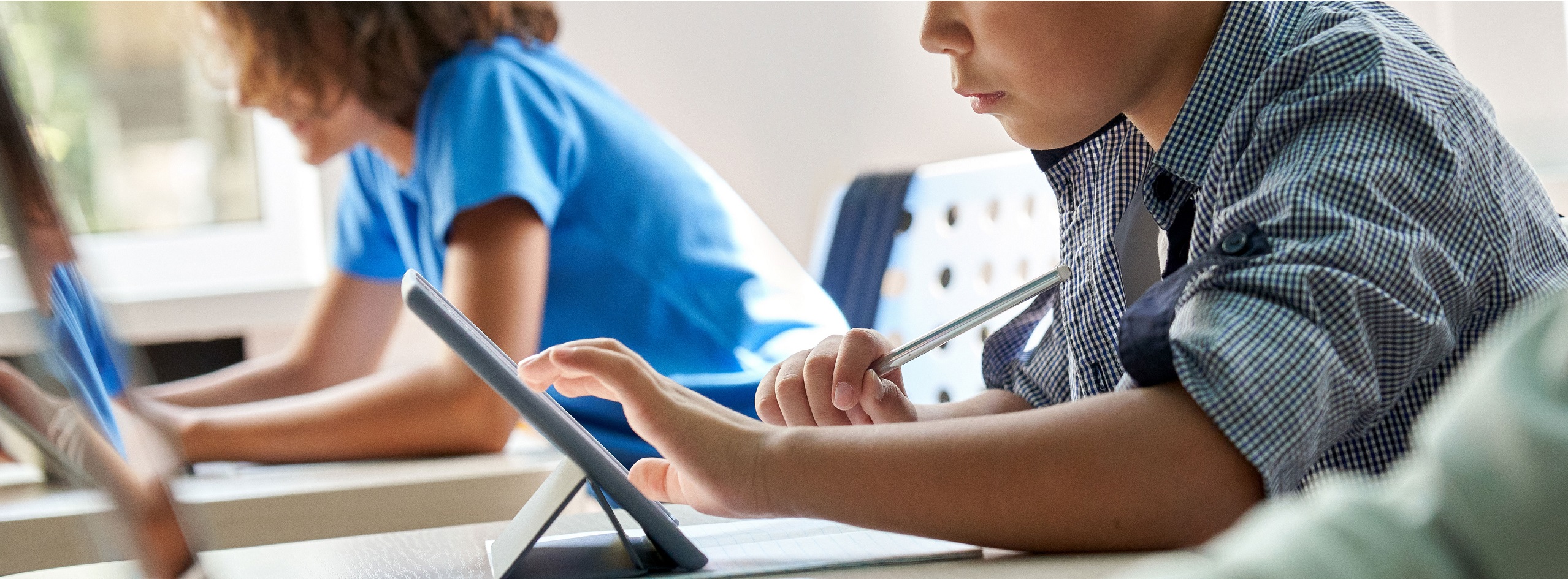 Zwei Kinder sitzen an einem Tisch und arbeiten mit Tablets. Ein Kind tippt auf dem Bildschirm, während es mit einem Stift auf Papier notiert. Beide konzentrieren sich auf ihre Aufgaben in einem modernen Lernumfeld.