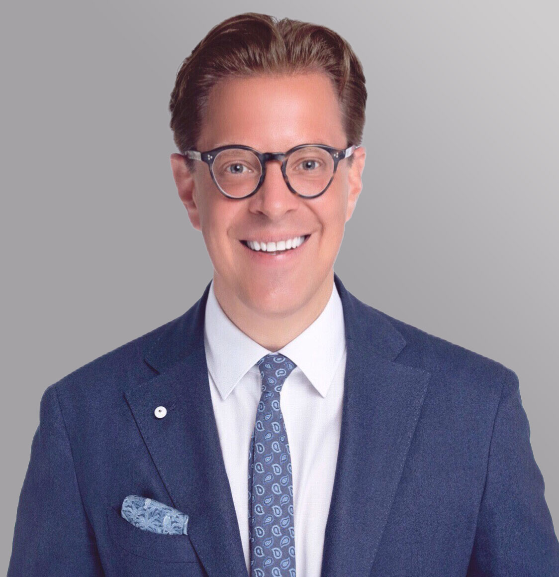 A smiling person with short, styled hair wears round glasses and a blazer with a pocket square. They are dressed in a collared shirt and tie, set against a solid light gray background. The expression conveys warmth and approachability.