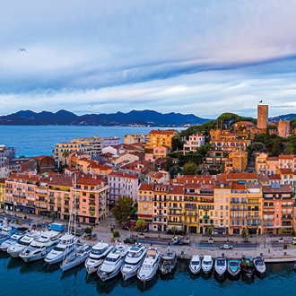 A scenic waterfront view showcases a vibrant town with colorful buildings along a marina. Various boats are docked in the water, while rolling hills and a cloudy sky form the backdrop. The image captures the charm of coastal life and picturesque architecture.