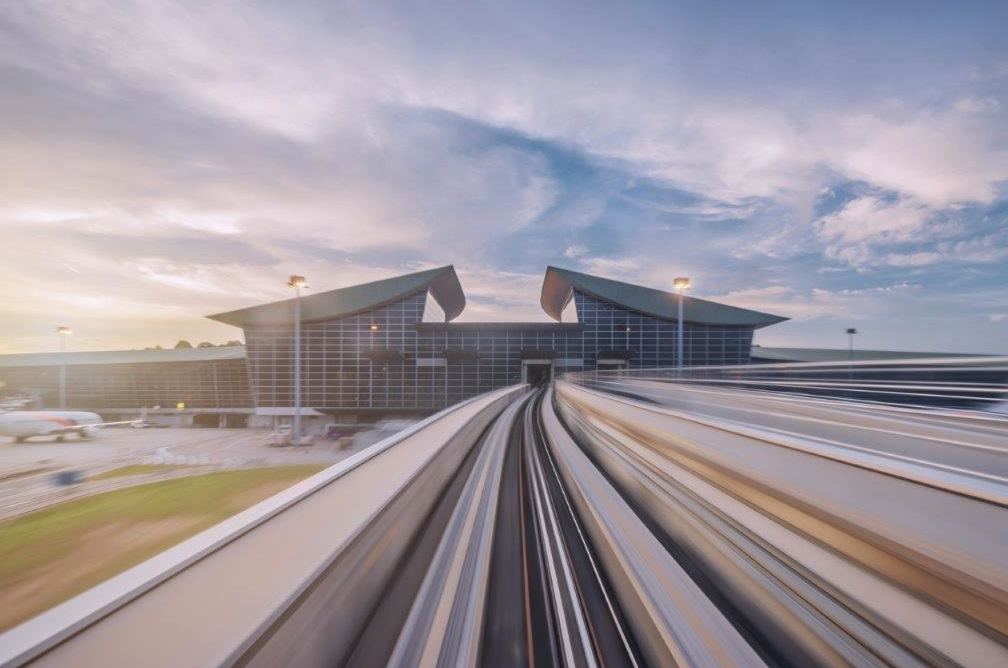 Ein moderner Flughafen mit einer großen, gläsernen Fassade und einem charakteristischen, geneigten Dach. Die Aufnahme vermittelt ein Gefühl von Bewegung, während eine elektrische Bahn in Richtung des Terminals unterwegs ist. Der Himmel zeigt sanfte Wolken und ein warmes Licht.