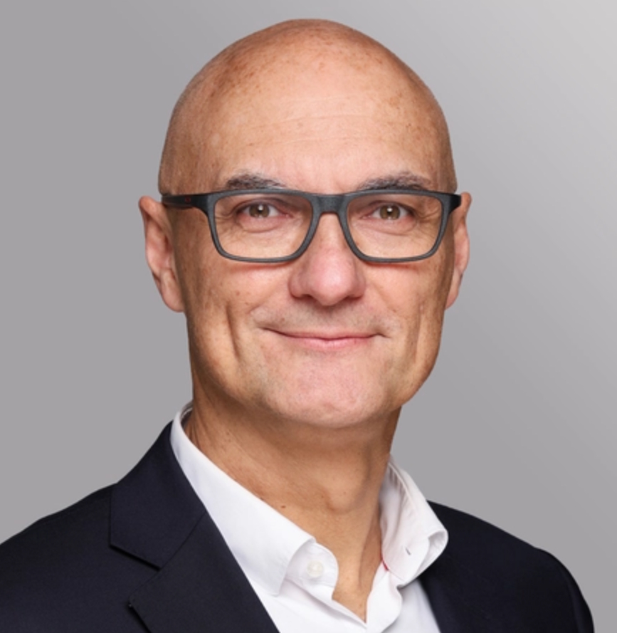 Ein mittelalterlicher Mann mit Glatze und Brille lächelt in die Kamera. Er trägt einen schwarzen Blazer über einem weißen Hemd. Der Hintergrund ist grau und unauffällig, um den Fokus auf das Gesicht zu legen.