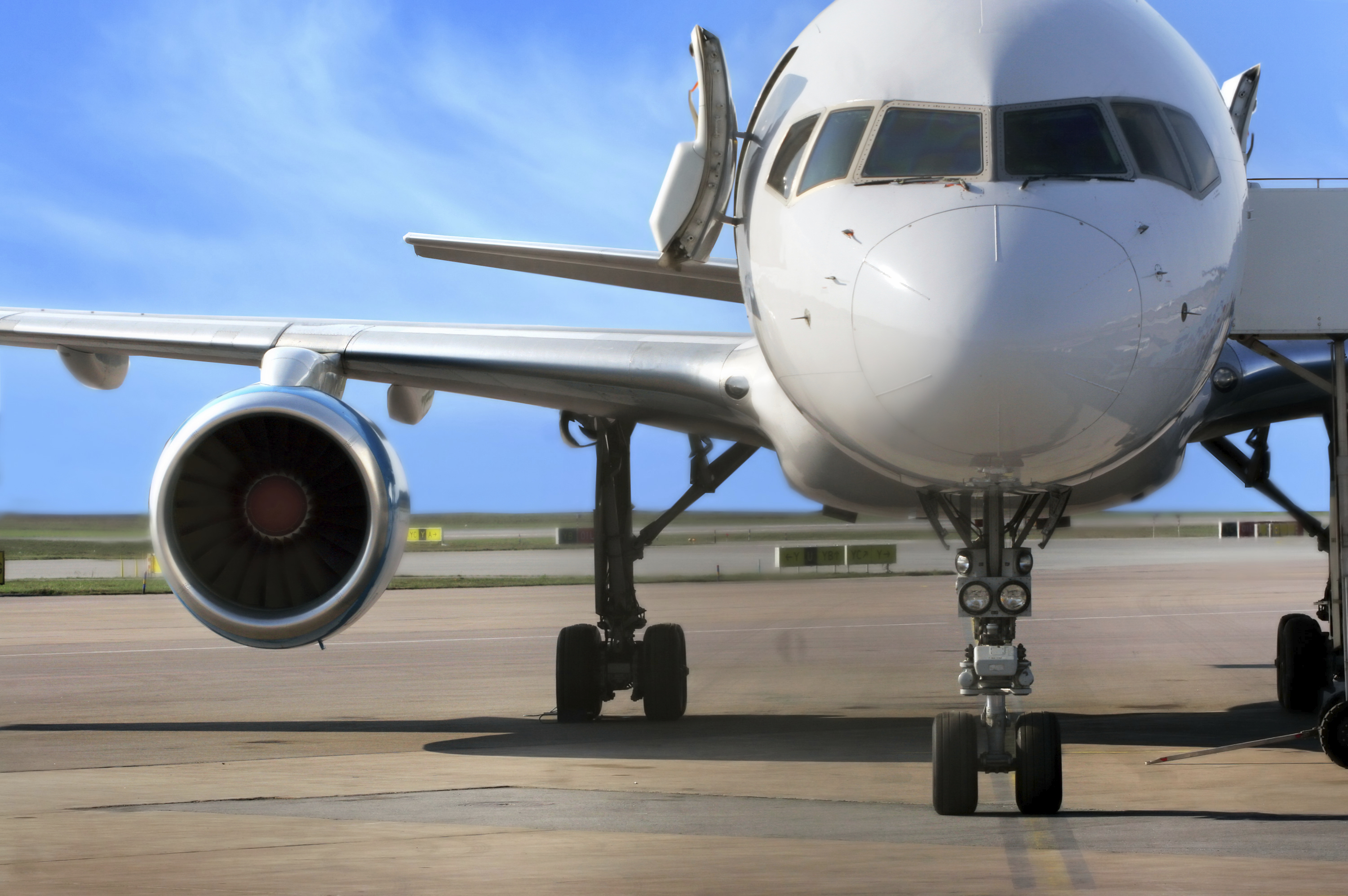 Ein Passagierflugzeug steht auf dem Flughafen vor einem blauen Himmel. Der Fokus liegt auf der Frontansicht des Flugzeugs, inklusive eines Triebwerks und der Fahrwerksräder. Die Tür des Flugzeugs ist geöffnet, was auf das Einsteigen von Passagieren hinweisen könnte.