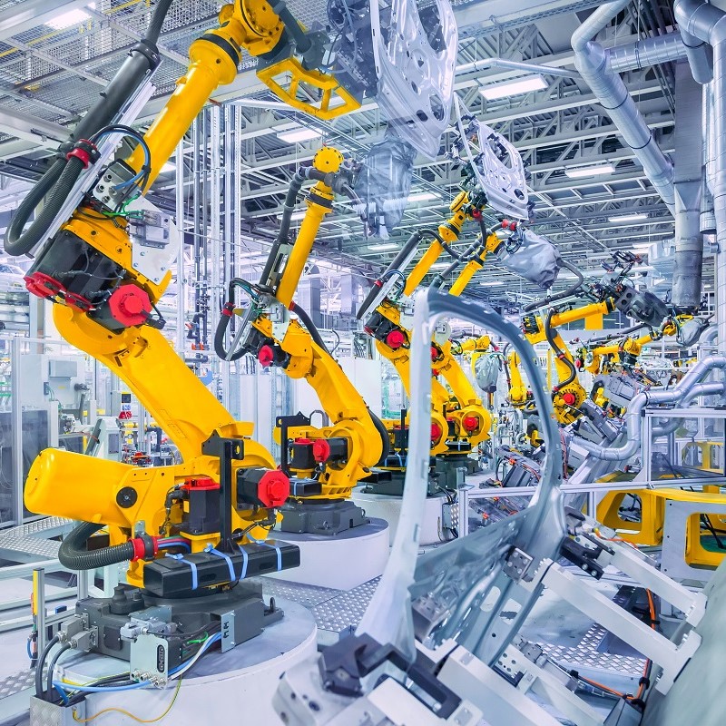 Eine moderne Fertigungsstraße mit mehreren gelben Industrierobotern, die Teile für Fahrzeuge montieren. Die Roboter sind auf runden Podesten platziert und arbeiten in einer hell erleuchteten, hochmodernen Umgebung.